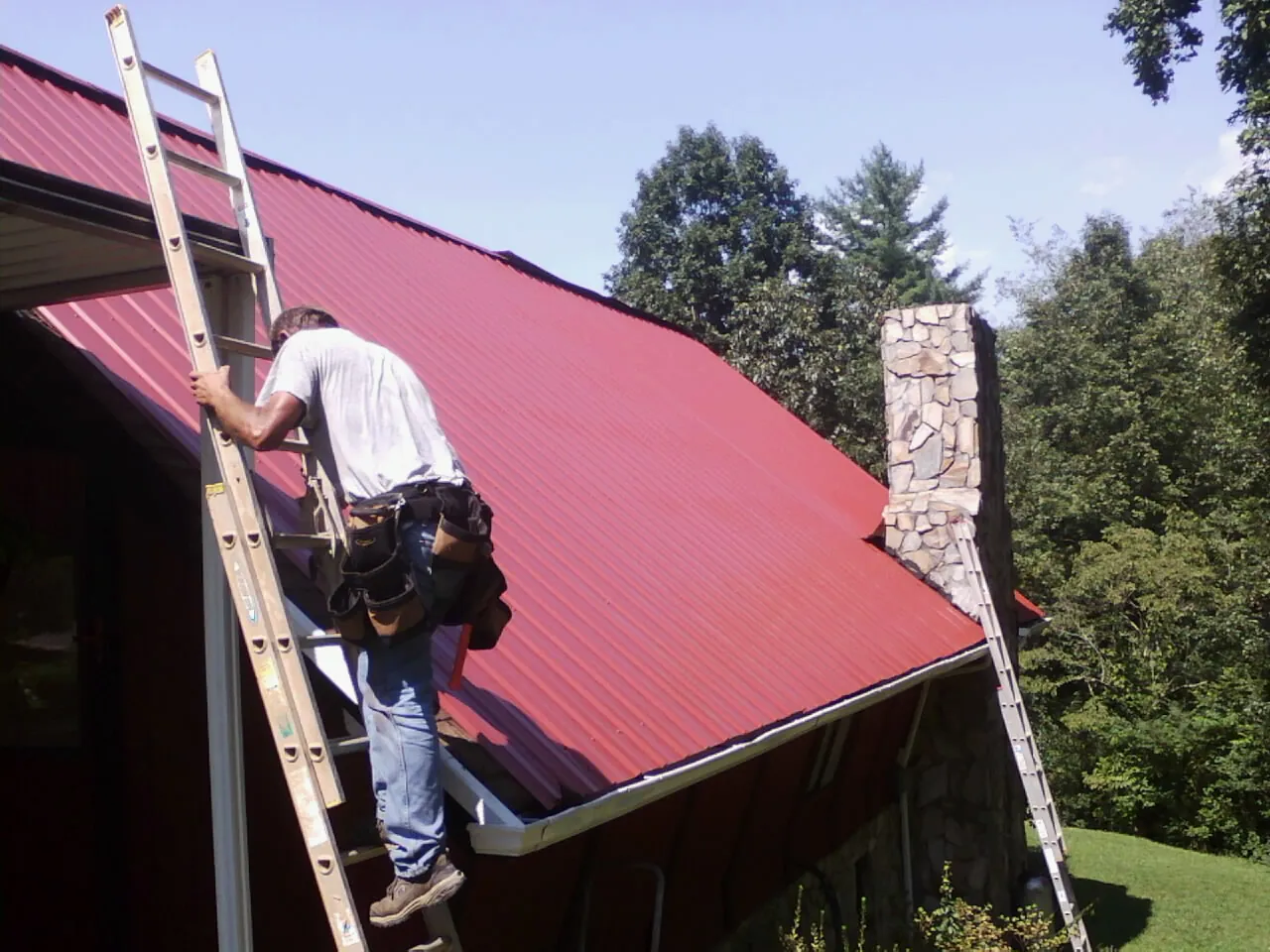 Roofer on a ladder beside a red metal roof and stone chimney — Soffit & Fascia Repair by Roofing Brothers in Purchase